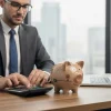 Мужчина в деловом костюме и очках, сидящий за рабочим столом, набирает что-то на калькуляторе. На столе стоит светло-коричневая керамическая копилка-свинья с нарисованными символами сбережений. Справа открытый ноутбук с финансовыми графиками на экране. На фоне — размытый городской пейзаж.