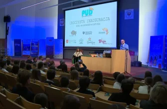 Церемония открытия 17-го академического года Подляского Детского Университета. На сцене — оратор за трибуной и женщина в академической мантии (ректор/декан), обращенные к полному залу учеников 4–6 классов, которые получили индексы. На экране: «UROCZYSTA INAUGURACJA ROKU AKADEMICKIEGO 2025/2026» и логотип PUD.