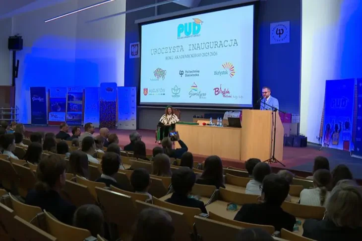 Церемония открытия 17-го академического года Подляского Детского Университета. На сцене — оратор за трибуной и женщина в академической мантии (ректор/декан), обращенные к полному залу учеников 4–6 классов, которые получили индексы. На экране: «UROCZYSTA INAUGURACJA ROKU AKADEMICKIEGO 2025/2026» и логотип PUD.