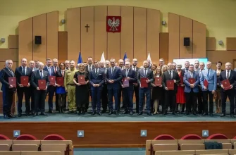 Большая группа высокопоставленных лиц, мужчин и женщин, стоящих на сцене в конференц-зале в Белостоке. Почти все держат в руках красные папки или документы. На заднем плане — Герб Польши (Орел) и флаги, а также деревянная стена с крестом.