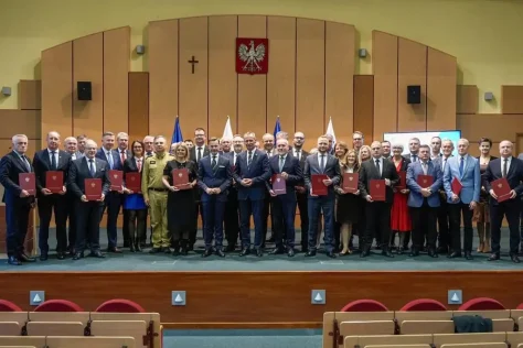 Большая группа высокопоставленных лиц, мужчин и женщин, стоящих на сцене в конференц-зале в Белостоке. Почти все держат в руках красные папки или документы. На заднем плане — Герб Польши (Орел) и флаги, а также деревянная стена с крестом.