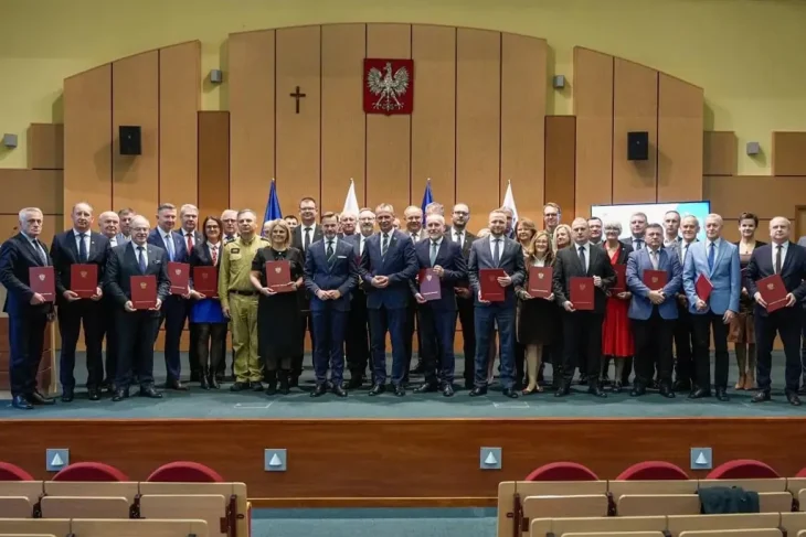 Большая группа высокопоставленных лиц, мужчин и женщин, стоящих на сцене в конференц-зале в Белостоке. Почти все держат в руках красные папки или документы. На заднем плане — Герб Польши (Орел) и флаги, а также деревянная стена с крестом.