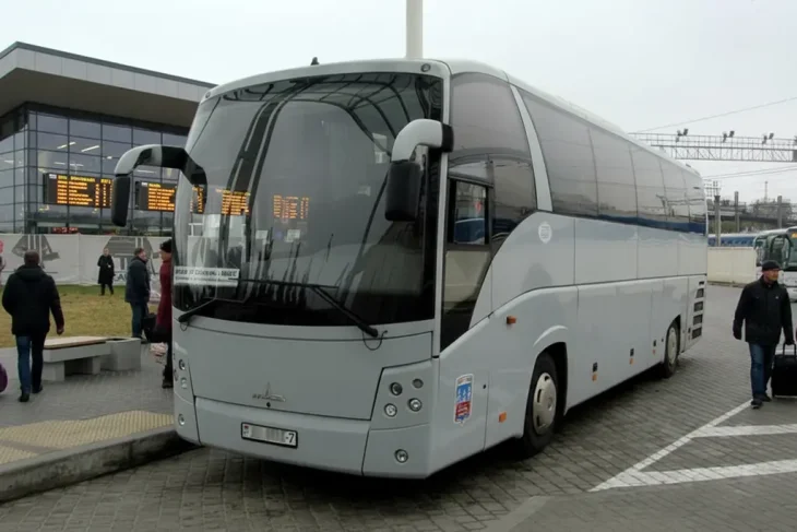 Светло-серый туристический автобус марки "МАЗ", принадлежащий ГП "Минсктранс" стоит на стоянке недалеко от стеклянного здания автовокзала.
