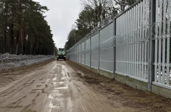 Физическая ограда на польско-беларуской границе, состоящая из высокого металлического забора с колючей проволокой, проходящая через густой лес. По грязной дороге едет военный или пограничный автомобиль.