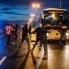 Ночная сцена конфликта на трассе S8 в Польше. Группа людей с закрытыми лицами и дубинками атакует туристический автобус "Madrid Travel". Прибывший полицейский с щитом пытается сдержать нападавших.