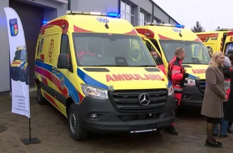 Два новые желто-красно-синие автомобиля скорой медицинской помощи Mercedes-Benz Sprinter с включенными синими проблесковыми маячками. На переднем плане — двое мужчин в красной униформе спасателя и женщина в бежевом пальто.