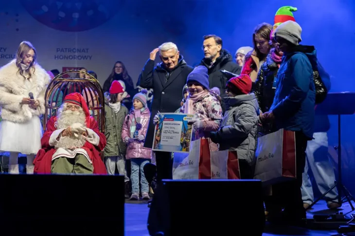 Сцена награждения или раздачи подарков: Санта-Клаус из Рованиеми сидит в кресле на сцене в Белостоке, а дети в зимней одежде получают призы и подарочные пакеты из рук организаторов.