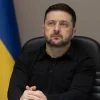Президент Украины Владимир Зеленский в черной рубашке сидит в кресле на фоне флага