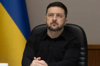 Президент Украины Владимир Зеленский в черной рубашке сидит в кресле на фоне флага