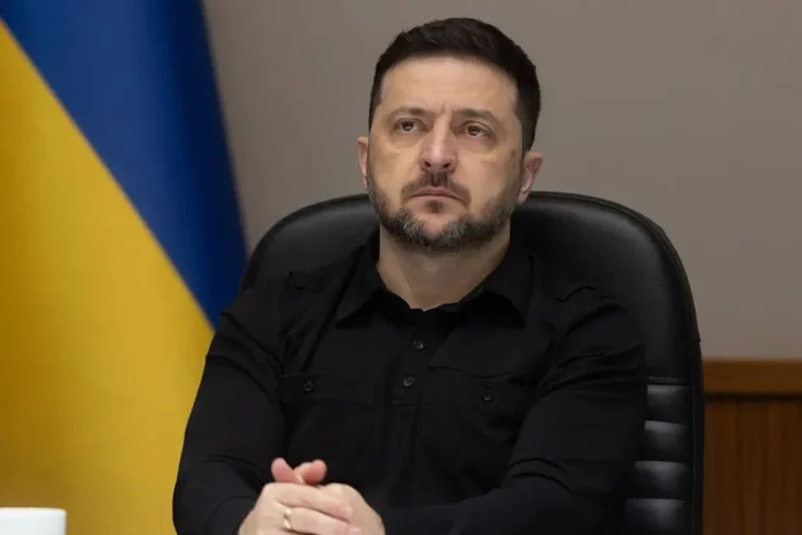 Президент Украины Владимир Зеленский в черной рубашке сидит в кресле на фоне флага